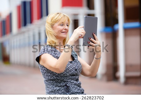 Happy smiling rich senior business woman 60-65 years old using ipad tablet pc computer reading ebook in city park