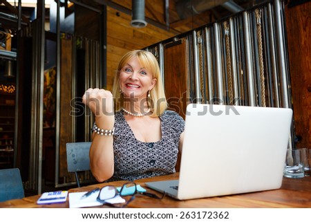 Smiling old senior blonde business woman 60-65 years, alone with laptop in summer cafe with gesture on face and raised hand up with fist