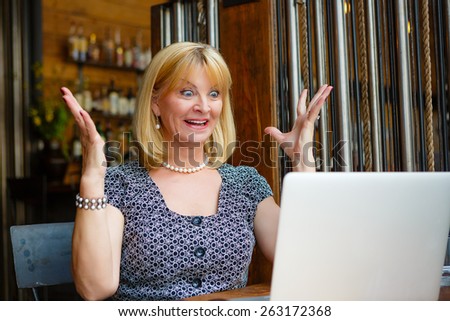 Old senior blonde business woman 60-65 years, alone with laptop in summer cafe with surprise and gesture on his face