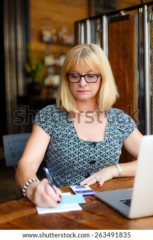 Old senior attractive business woman in the glasses sitting at a cafe city with a laptop and calculator, write notes