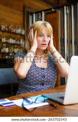 Old senior business woman with scared emotion on face, looking to computer laptop sitting in summer cafe place