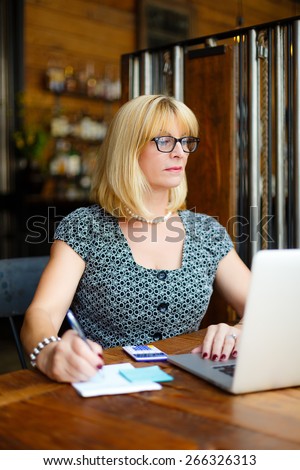 Old senior blonde business woman in the glasses sitting at a cafe city with a laptop and calculator, writes notes