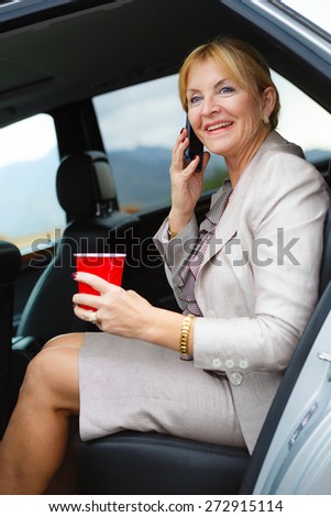 Happy and smile old senior woman 60-65 years with white teeth talking by mobile cell phone in the back of a car and holding plastic glass in hand.