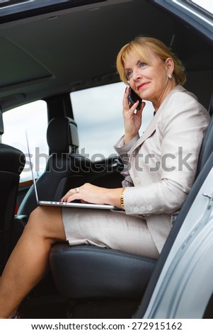 Old senior business woman 60-65 years talking by mobile cell phone in the back of a car and holding her Laptop Computer pc in lap