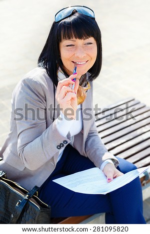 Stylish smile brunette pretty woman writing a letter and thinking with Pen in mouth, sitting on a park bench, sunny day, good weather, city street, human emotion, facial expression, spring mood