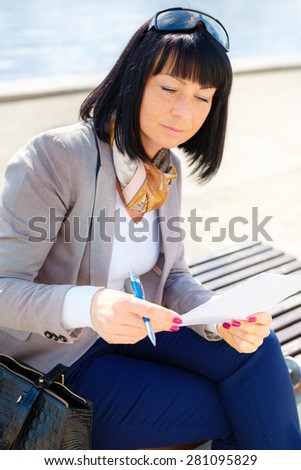 Stylish brunette pretty woman writing a letter and thinking with Pen in hand, sitting on a park bench, sunny day, good weather, city street, human emotion, facial expression, spring mood