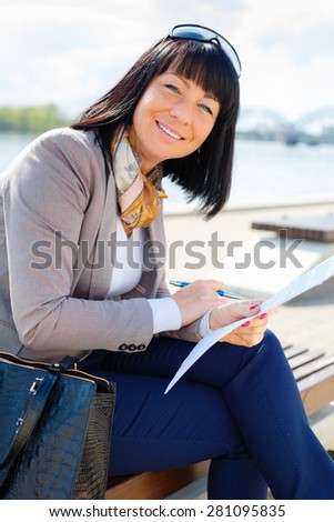 Stylish smile brunette pretty woman writing a letter and thinking with Pen in hand, sitting on a park bench, sunny day, good weather, city street, human emotion, facial expression, spring mood