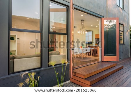 Double open french doors leading into contemporary home with wooden terrace, open floor plan, greenery at night. Large window showing modern interior at twilight.