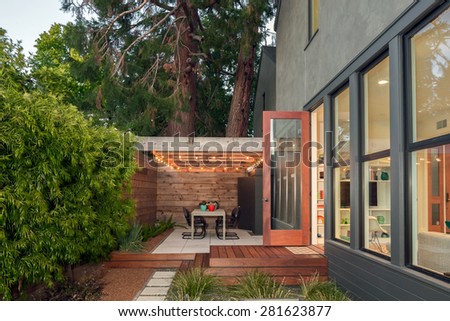 Open doors leading into contemporary home with wooden terrace and illuminated seating arrangement, open floor plan and greenery at night. Large window showing modern interior at twilight.