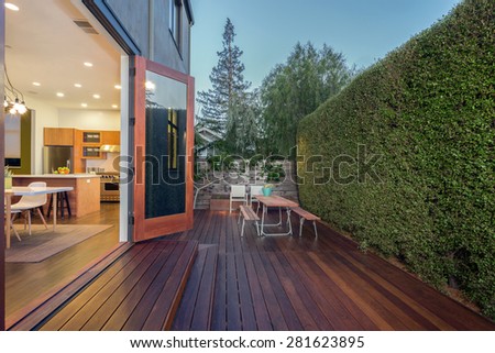Open french doors leading into contemporary home with wooden terrace, open floor plan, greenery at night. Large window showing modern interior at twilight.