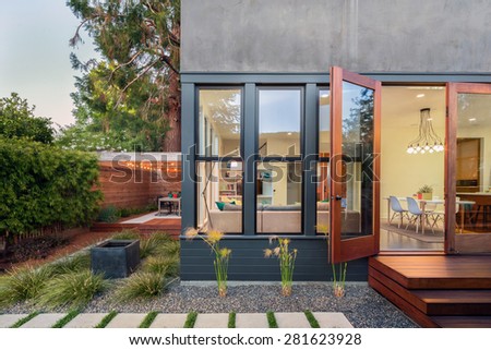 Open french doors leading into contemporary home with wooden terrace in Zen Garden style, open floor plan and greenery at night. Large window showing modern interior at twilight.