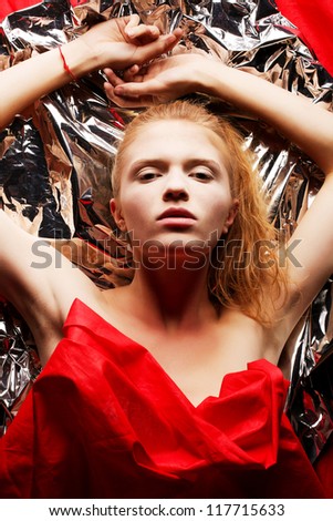 Arty portrait of a fashionable red-haired model in red with hands up over wrinkled silver foil background. Studio shot