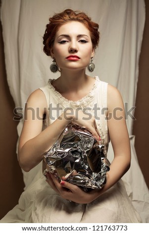 Arty portrait of a fashionable queen-like ginger model holding silver foil sphere over white curtain background. Studio shot