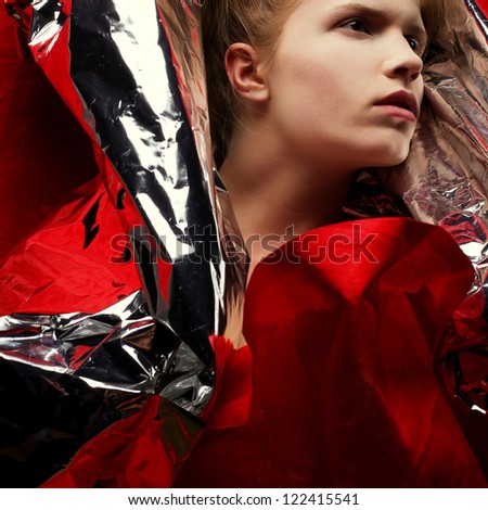 Arty portrait of a fashionable red-haired model in red with silver foil cape over red curtain background. Close-up. Studio shot