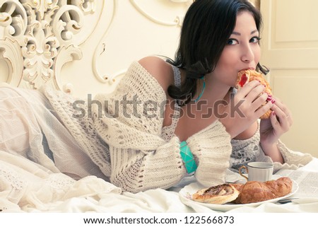 Portrait of a young beautiful woman eating her croissant with strawberry jam in a vintage bedroom. Indoor shot