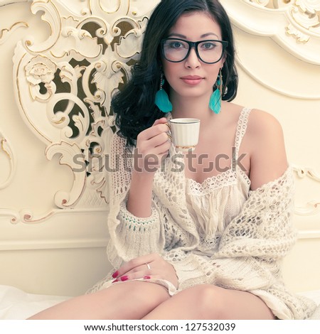 Portrait of a beautiful smiling girl in trendy glasses drinking tea or coffee in her vintage bedroom. Cup of hot beverage. Indoor shot