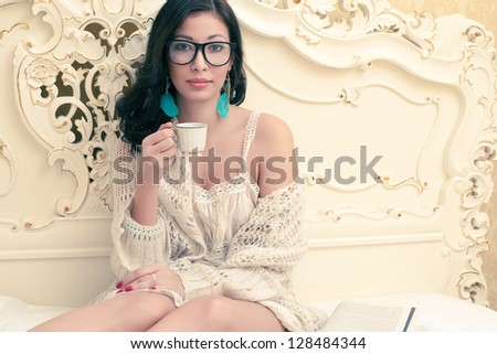 Portrait of a beautiful smiling girl in trendy glasses drinking tea or coffee in her vintage bedroom. Cup of hot beverage. Indoor shot