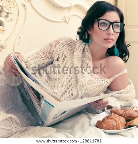 Portrait of a young beautiful brunette in a trendy glasses having breakfast (tea (coffee), croissants) and reading her morning newspaper in a vintage bedroom. Indoor shot