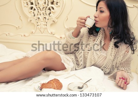 Portrait of a young beautiful brunette having breakfast (tea (coffee), croissants) and posing on a bed in her vintage bedroom. Copy-space. Indoor shot