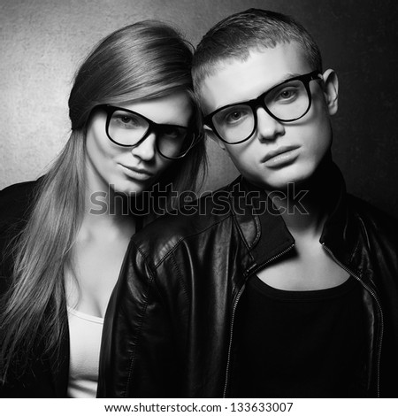 Portrait of gorgeous fashion twins in black clothes wearing trendy glasses and posing over gray background together. Perfect hair. Natural make-up. Perfect skin. Hipster style. Monochrome studio shot.