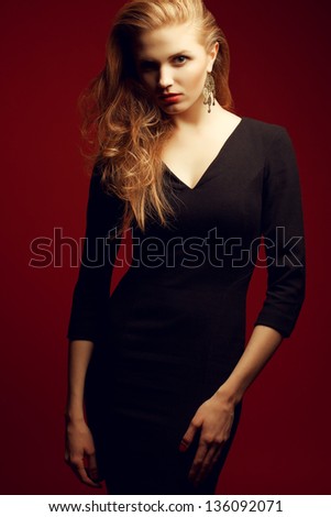 Portrait of a red-haired (ginger) fashionable model in a black classic cocktail dress posing over red background. Seductive glance. Vogue style. Studio shot