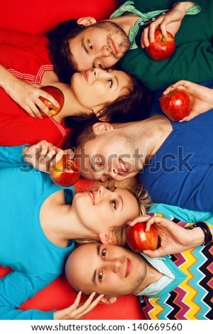 Portrait of five stylish close friends hugging and lying over red background. Guys having fun and holding red apples. Healthy food concept. Hipster style. Studio shot