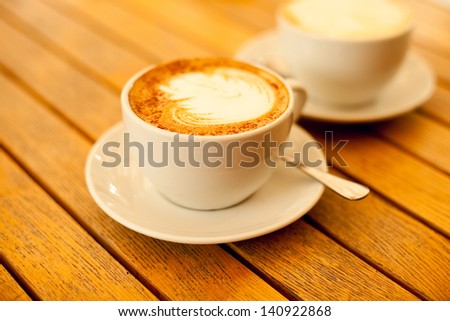 Latte art concept. Two cups with cappuccino (hot coffee with milk foam) and canella (cinnamon) on wooden table at street cafe (coffe bar). Vintage style. Close up. Outdoor shot.