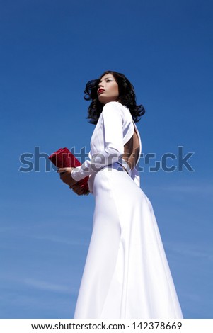 Portrait of a beautiful woman (fashion model) posing in elegant white atlas cocktail dress with red clutch in her hands over blue sky background. Copy-space. Outdoor shot