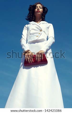 Portrait of a beautiful woman (fashion model) posing in elegant white atlas cocktail dress with red leather clutch in her hands over blue sky background. Outdoor shot