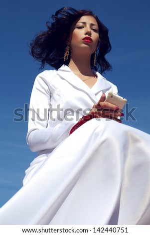 Portrait of a beautiful woman posing in elegant white atlas cocktail dress with red leather clutch holding mobile phone. Luxurious golden accessories (ring, earrings). Outdoor shot
