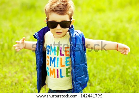 Stylish Baby Boy With Ginger (Red) Hair In Trendy Sunglasses And Blue Jacket Standing In The Park &Amp; Trying To Hug Somebody. Hipster Style. Sunny Weather. Smile Word Printed On T-Shirt. Outdoor Shot