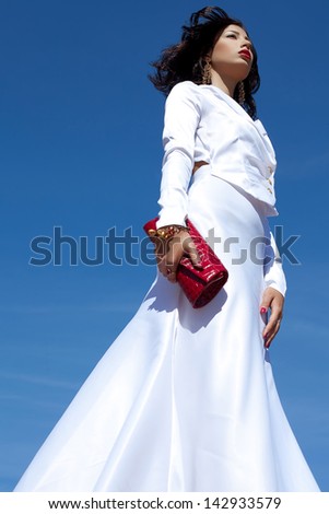 Portrait of a beautiful woman posing in elegant white atlas cocktail dress with red leather clutch in her hands over blue sky background. Luxurious golden accessories (bracelet, earrings). Copyspace