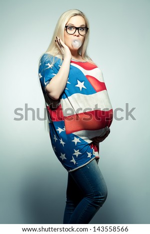 American Mom Concept: Surpised young pregnant woman in american flag like dress and trendy glasses chewing bubble gum over gray background. Hipster style. Studio shot