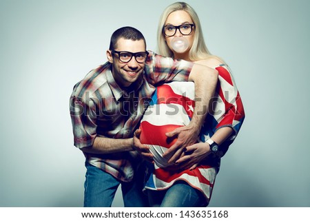 Happy american family (pregnancy) concept: portrait of two funny hipsters (husband and wife) in trendy glasses and clothes playing with their bubble gum. Copy-space. Studio shot