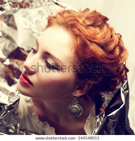 Arty portrait of a fashionable queen-like red-haired (ginger) model with silver foil cape posing over white curtain background. Vintage (classic, retro) style. Close up. Studio shot