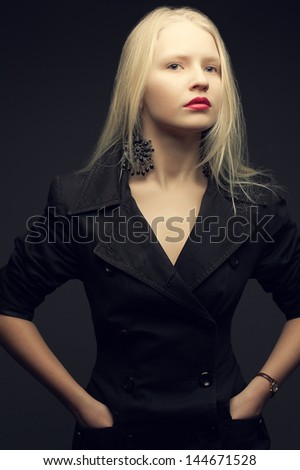 Portrait of a beautiful fashionable model with natural blond hair and great make-up in trendy black coat posing over dark gray background. Hands in pockets. Studio shot