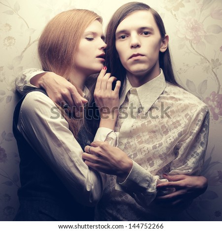 Portrait of beautiful long haired people in vintage style: handsome boy with brown hair and whispering gorgeous red-haired girl posing together. Studio shot