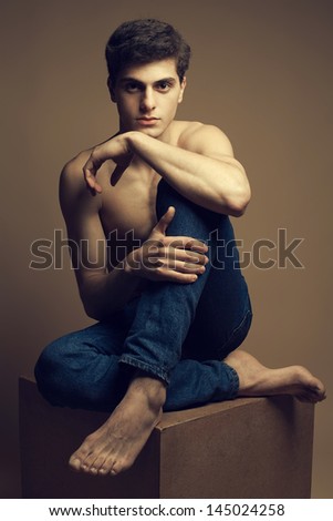 Blue jeans concept. Beautiful (handsome) muscular young man in jeans with great body sitting on a wooden cube and posing over wooden background. Fashion studio portrait