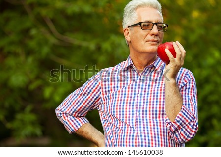 Healthcare concept. Portrait of a smiling mature (old) man in casual shirt and stylish glasses holding red apple and posing in the park. Copy-space. Outdoor shot