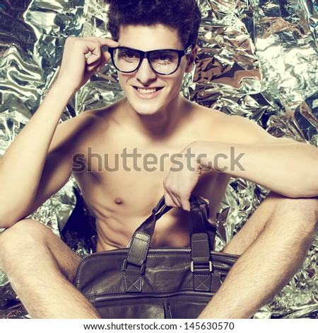 Smiling beautiful (handsome) muscular male model with nice body in trendy glasses posing over wrinkled silver foil background with stylish leather bag. Hipster (vintage) style. Fashion studio portrait