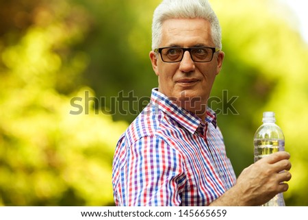 Portrait of a smiling happy mature (old) man in trendy casual shirt and glasses posing in the park and drinking water. Sunny summer day. Copy-space. Close up. Outdoor shot
