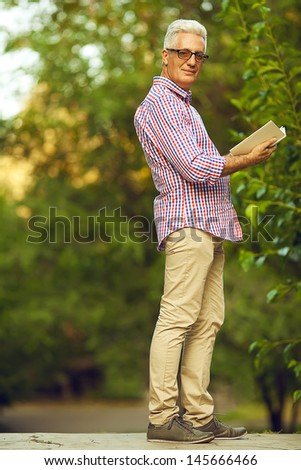 Successful man concept. Portrait of a smiling happy mature (old) man in trendy casual shirt & glasses looking at camera and trying to read book in the park. Sunny summer day. Copy-space. Outdoor shot