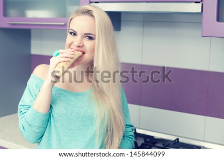 Healthy pregnancy concept. Portrait of a lovely and funny young pregnant woman eating her brioche (bread)  in her modern hi-tech kitchen. Trendy clothing on girl. Copy-space. Indoor shot