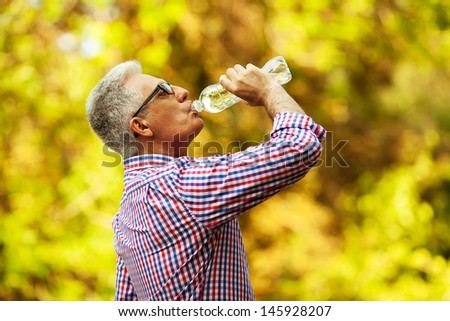 Portrait of a mature (old) man in trendy casual shirt and glasses posing in the park and drinking water. Sunny summer day. Copy-space. Outdoor shot