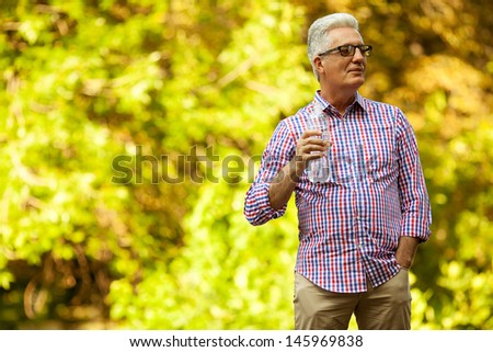 Portrait of a mature (old) man in trendy casual shirt and glasses posing in the park and drinking water. Sunny summer day. Copy-space. Outdoor shot