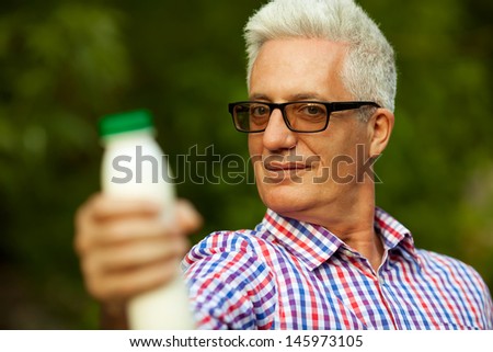 Healthy food concept. Portrait of a mature (old) man in trendy casual shirt and glasses posing in the park and holding a bottle with milk. Sunny summer day. Close up. Copy-space. Outdoor shot