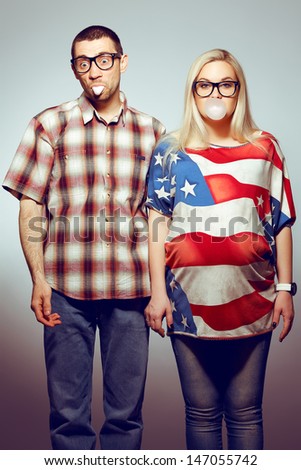 Funny pregnancy concept: portrait of two hipsters (husband and wife) in trendy glasses and clothes playing with their bubble gums. Studio shot