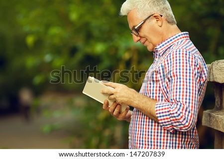 Successful man concept. Profile portrait of a smiling happy mature (old) man in trendy casual shirt & glasses reading book in the park. Sunny summer day. Copy-space. Outdoor shot