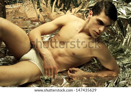 Beautiful (handsome) muscular male model with nice abs in sitting and posing over wrinkled silver foil background. Perfect skin and body. White underwear. Vogue style. Fashion studio portrait