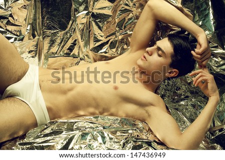 Beautiful (handsome) muscular male model with nice abs in sitting and posing over wrinkled silver foil background. Perfect skin and body. White underwear. Vogue style. Fashion studio portrait